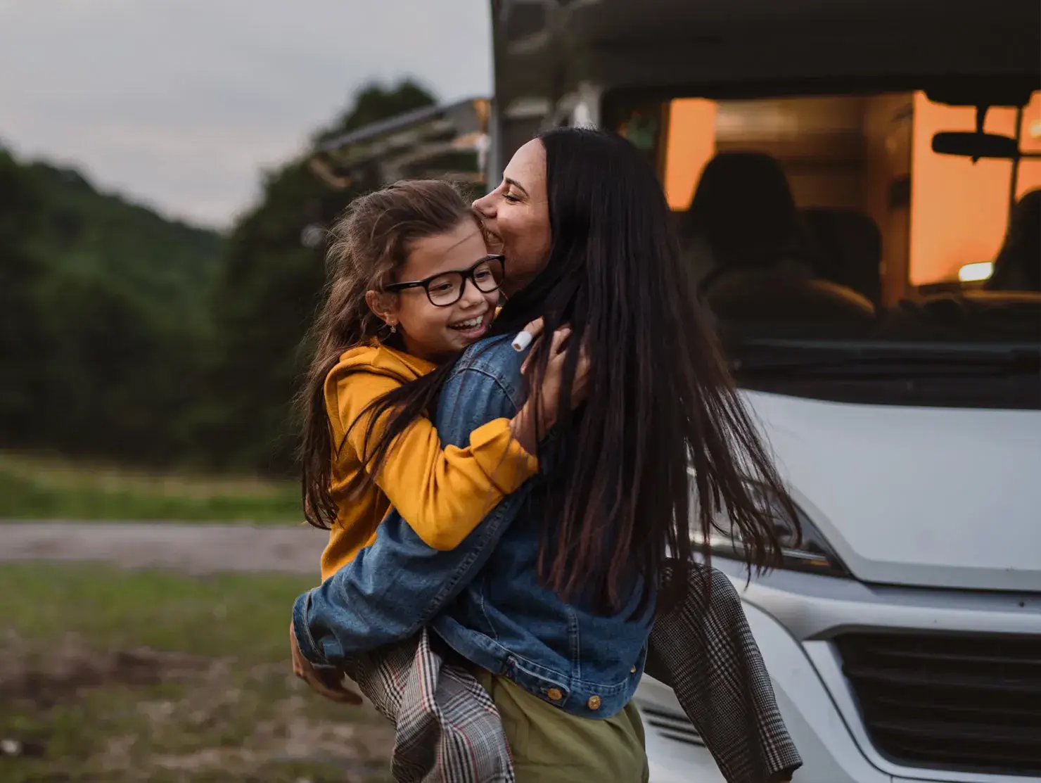 Mother lifting and hugging her smiling daughter in front of a caravan at dusk, for the CIL Weekends campaign highlighting family joy on the road.