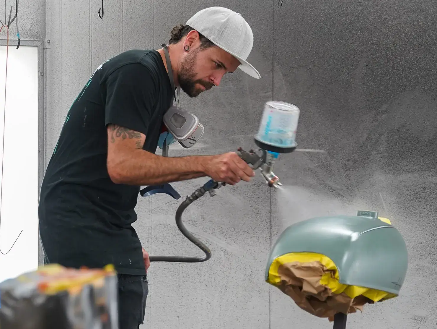 A custom painter carefully sprays a motorbike tank in a workshop, as part of the Shannons Dream Bike Build YouTube Series.
