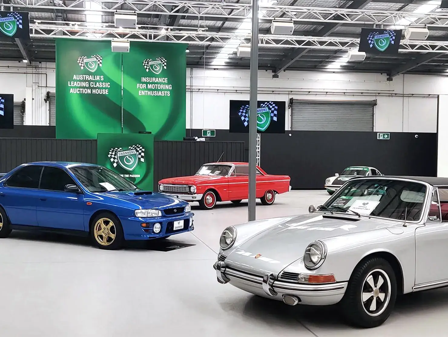 Shannons auction showroom with classic cars including a Porsche and Subaru, staged beneath branding banners for motoring enthusiasts.