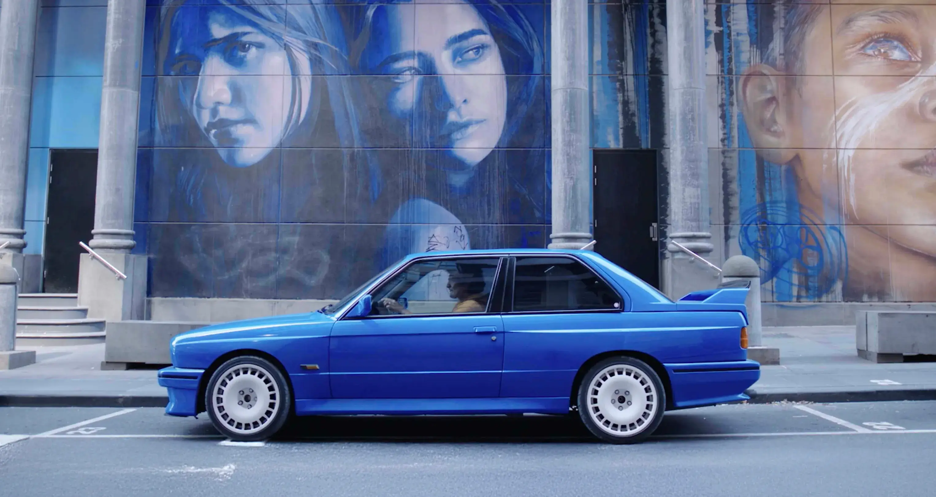 Blue BMW E30 M3 parked in front of a large street mural featuring portraits, part of the Shannons 'Share the Passion' campaign for under-40s. 