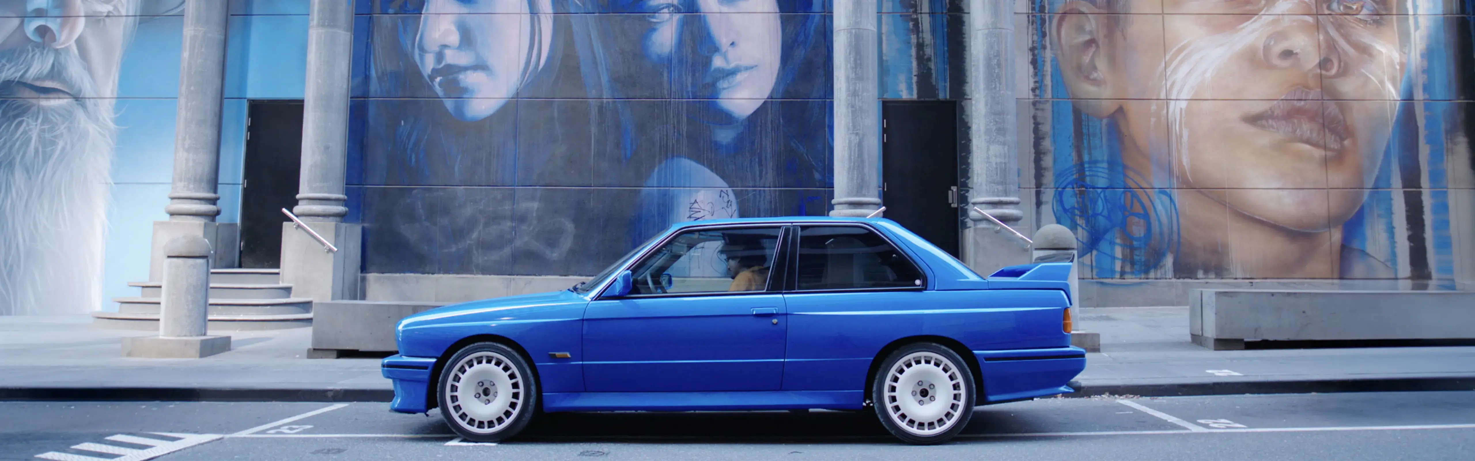 Blue BMW E30 M3 parked in front of a large street mural featuring portraits, part of the Shannons 'Share the Passion' campaign for under-40s.