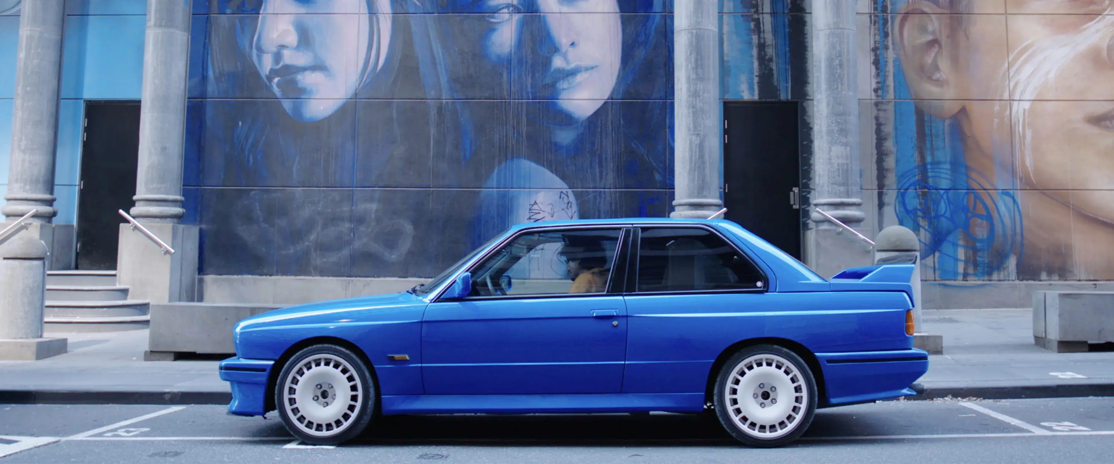 Blue BMW E30 M3 parked in front of a large street mural featuring portraits, part of the Shannons 'Share the Passion' campaign for under-40s.