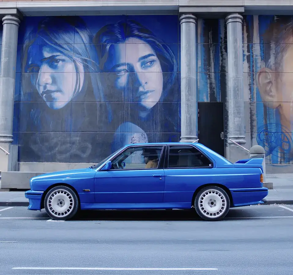 Blue BMW E30 M3 parked in front of a large street mural featuring portraits, part of the Shannons 'Share the Passion' campaign for under-40s.