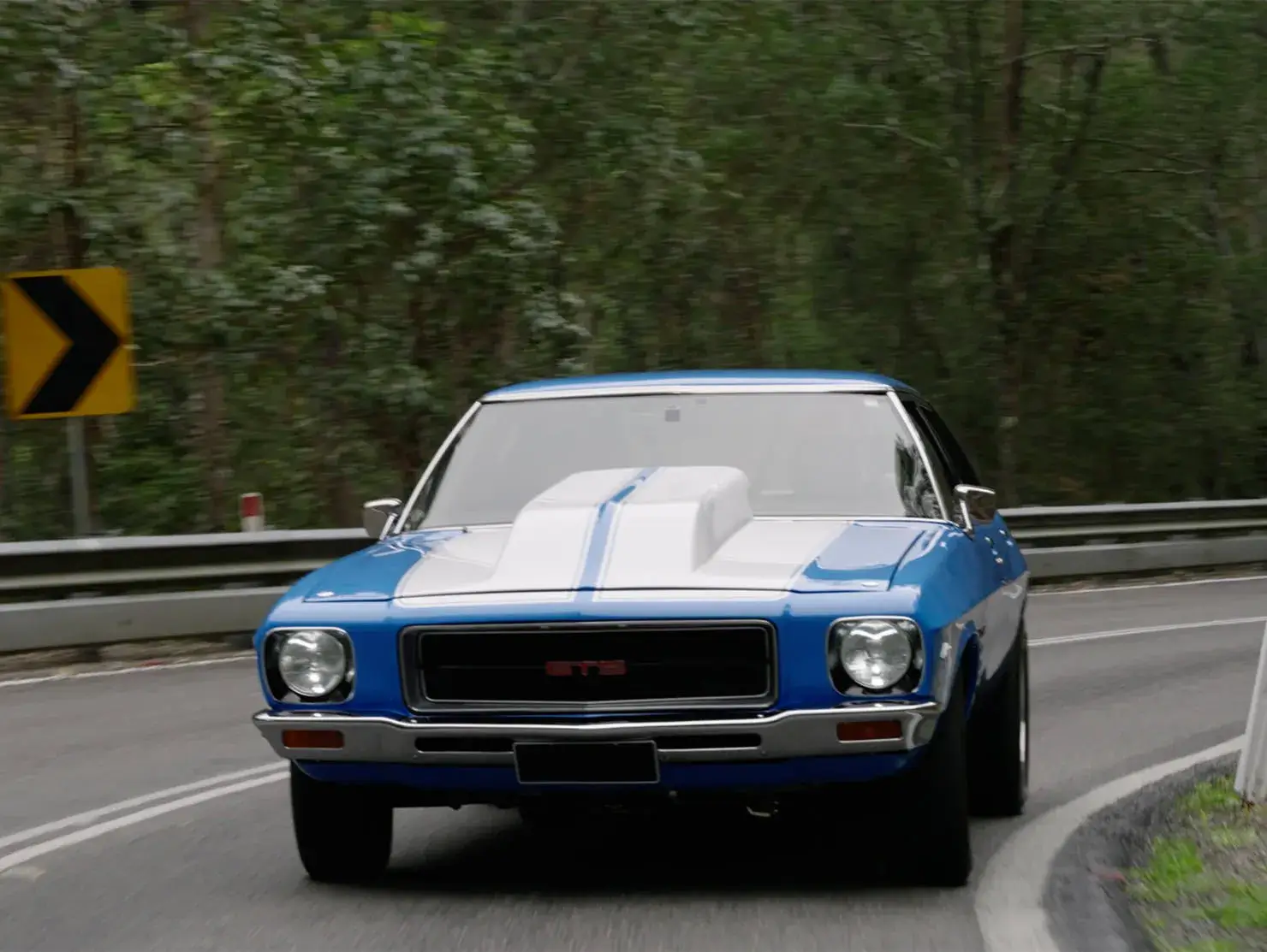 Classic blue Holden GTS with white racing stripes driving on a winding road, part of the Shannons 'Share the Passion' campaign for under-40s.