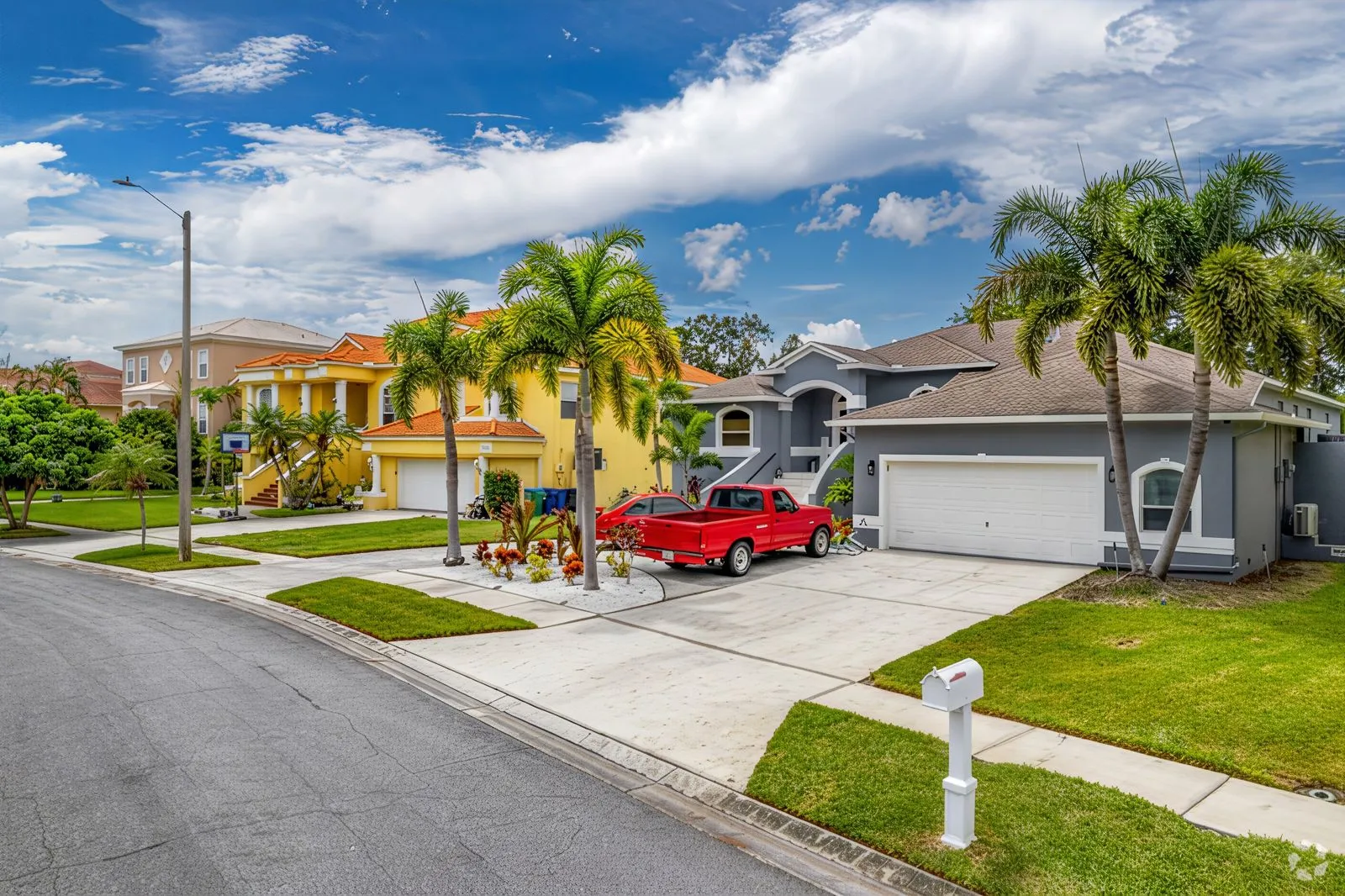 A suburban street in Seminole boasts neatly maintained houses featuring vibrant colors and palm trees. A red truck from is parked in the driveway of a gray house with a white garage door. The sky above is blue with scattered clouds, and a white mailbox stands near the sidewalk.