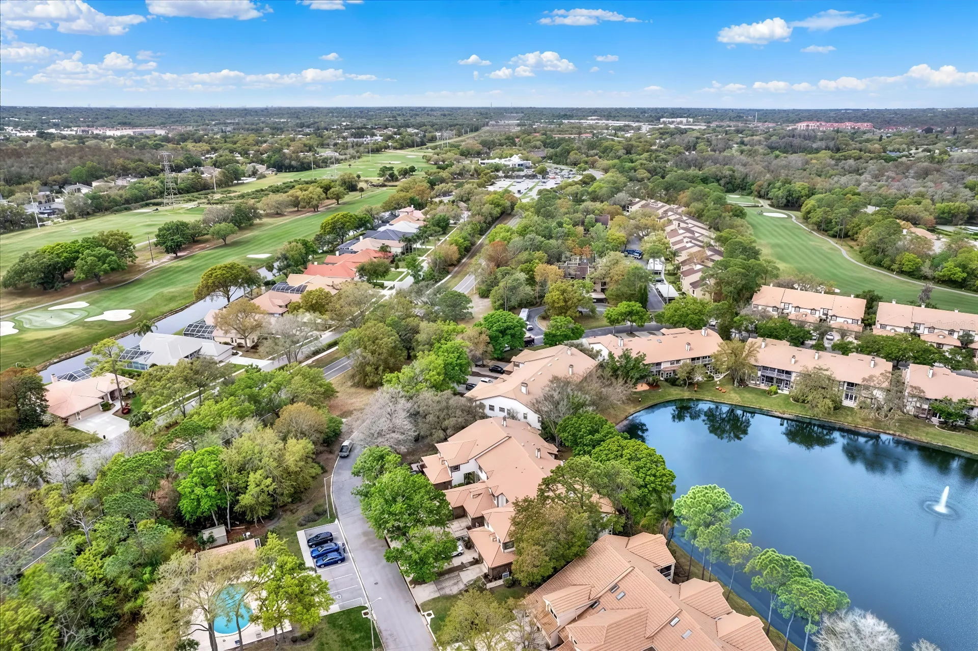 Aerial view of a suburban neighborhood with beige-roofed houses surrounded by a lush landscape. There is a clear pond with a fountain in the foreground, a golf course on the left, and a mix of trees and greenery throughout.