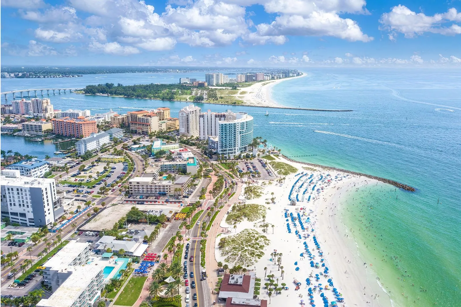 Aerial view of Clearwater, a vibrant beach city with crowded beaches lined with umbrellas, high-rise buildings, and a curving coastline. The turquoise waters stretch out to the horizon under a partly cloudy sky. A bridge connects the city to a green, tree-filled area.