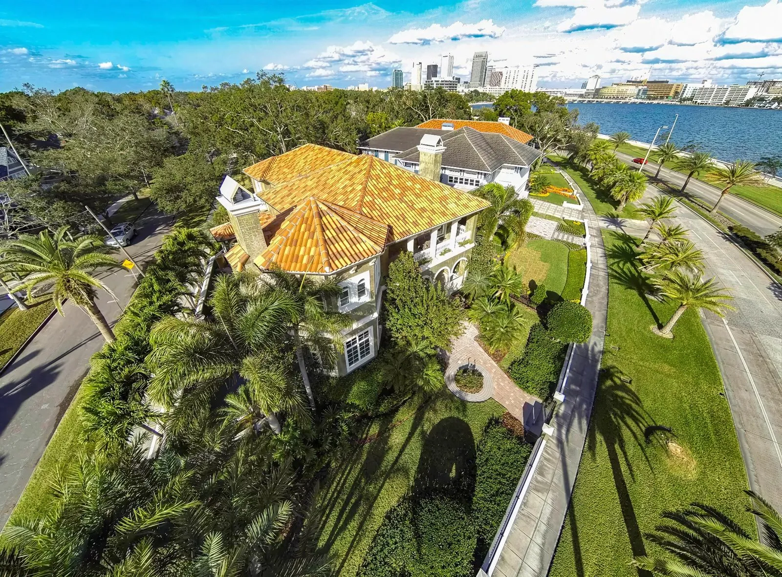 Aerial view of a luxurious mansion with a red-tiled roof surrounded by palm trees and greenery. Located near water in Tampa, the property features a well-maintained yard, stone pathway, and scenic city backdrop with a bright, cloudy sky