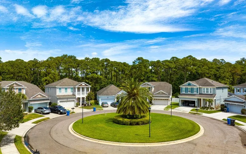 Aerial view of a suburban cul-de-sac in Town N’ Country, with five houses featuring driveways with parked cars and well-maintained lawns. A circular grassy island with a large palm tree graces the center, bordered by trees under a partly cloudy sky.