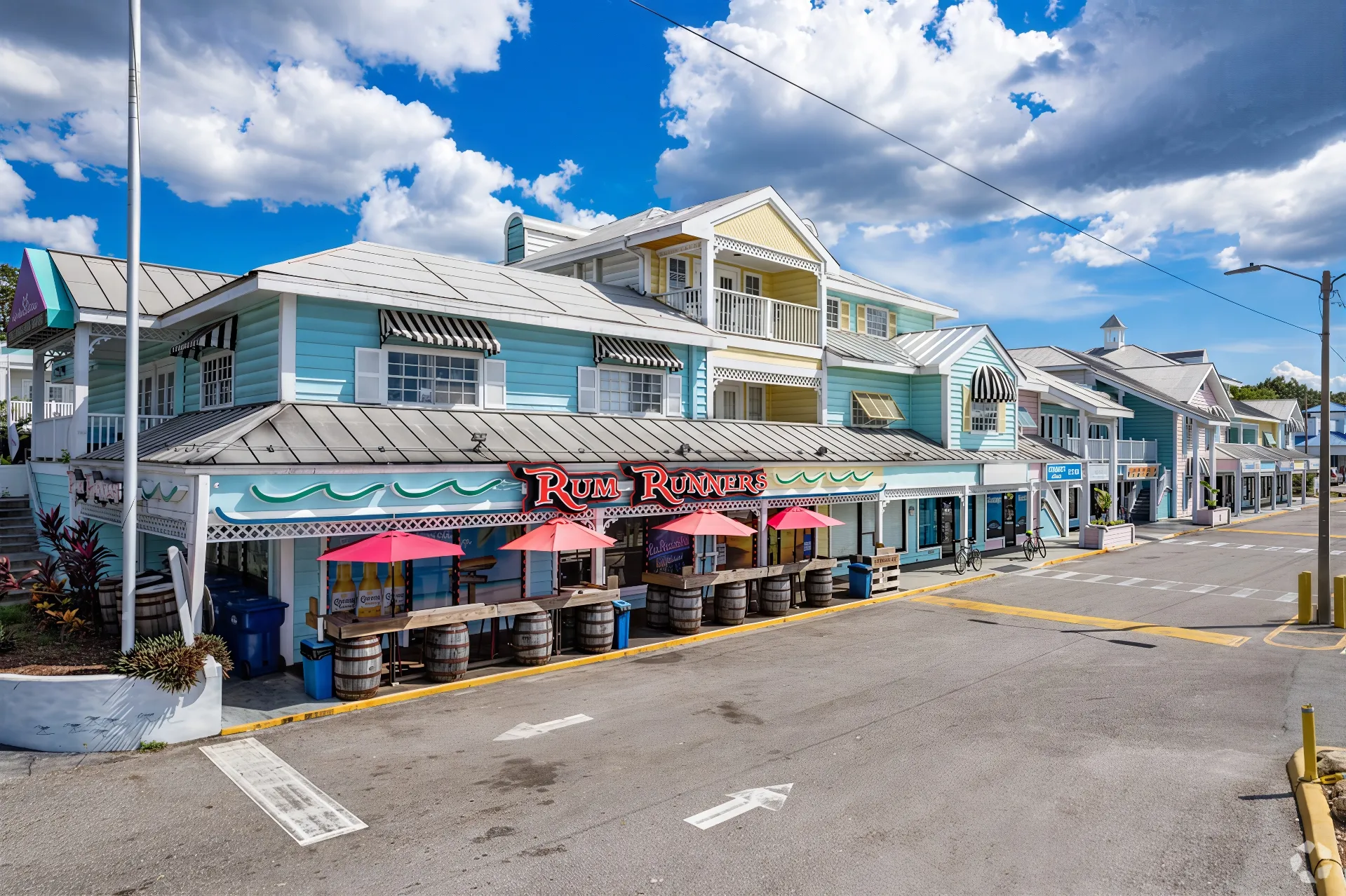 A colorful seaside two-story building houses the "Rum Runner" restaurant in Palm Harbor. The facade is adorned in pastel shades of blue and green, with outdoor seating under pink umbrellas. With a bright sky and fluffy clouds as the backdrop, this lively business feels welcoming and inviting.