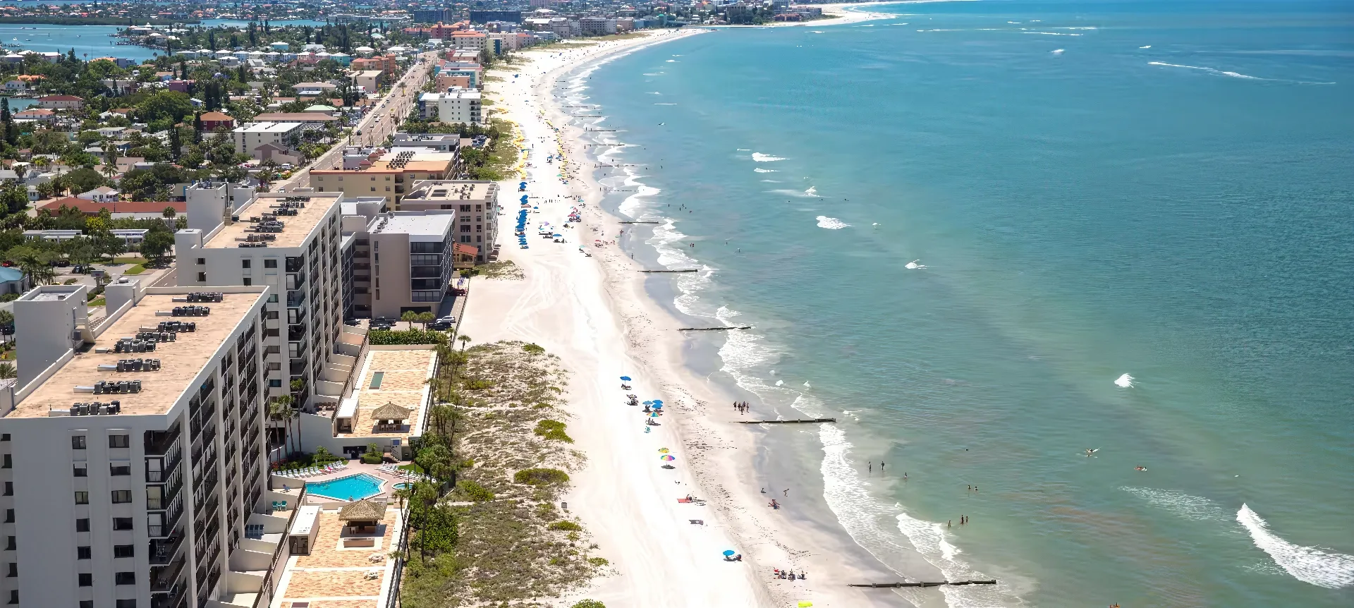 Aerial view of Madeira Beach lined with colorful umbrellas and buildings on the left side. The clear blue ocean is on the right, with gentle waves meeting the shore. The beach curves along the coastline, showcasing a town in the distance under a blue sky