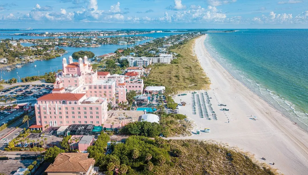 Aerial view of St. Pete Beach showcasing a large, pastel-pink building with Mediterranean architecture near a long, sandy beach. The shoreline hosts lined-up beach chairs and umbrellas, while nearby water bodies and residential buildings stretch out in the distance.