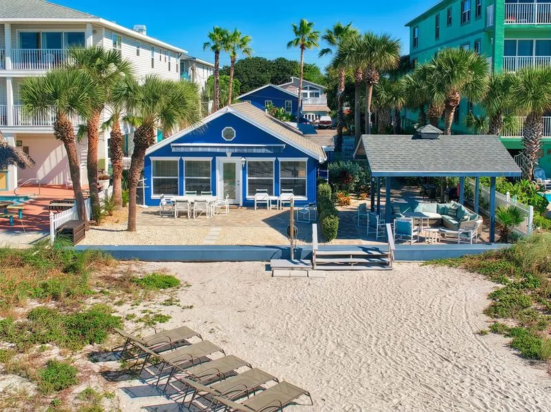 A beachfront property in Indian Rocks features a blue house with white trim, flanked by palm trees and adjacent buildings. In front of the house, there is a sandy area with lounge chairs and a gazebo furnished with outdoor seating.