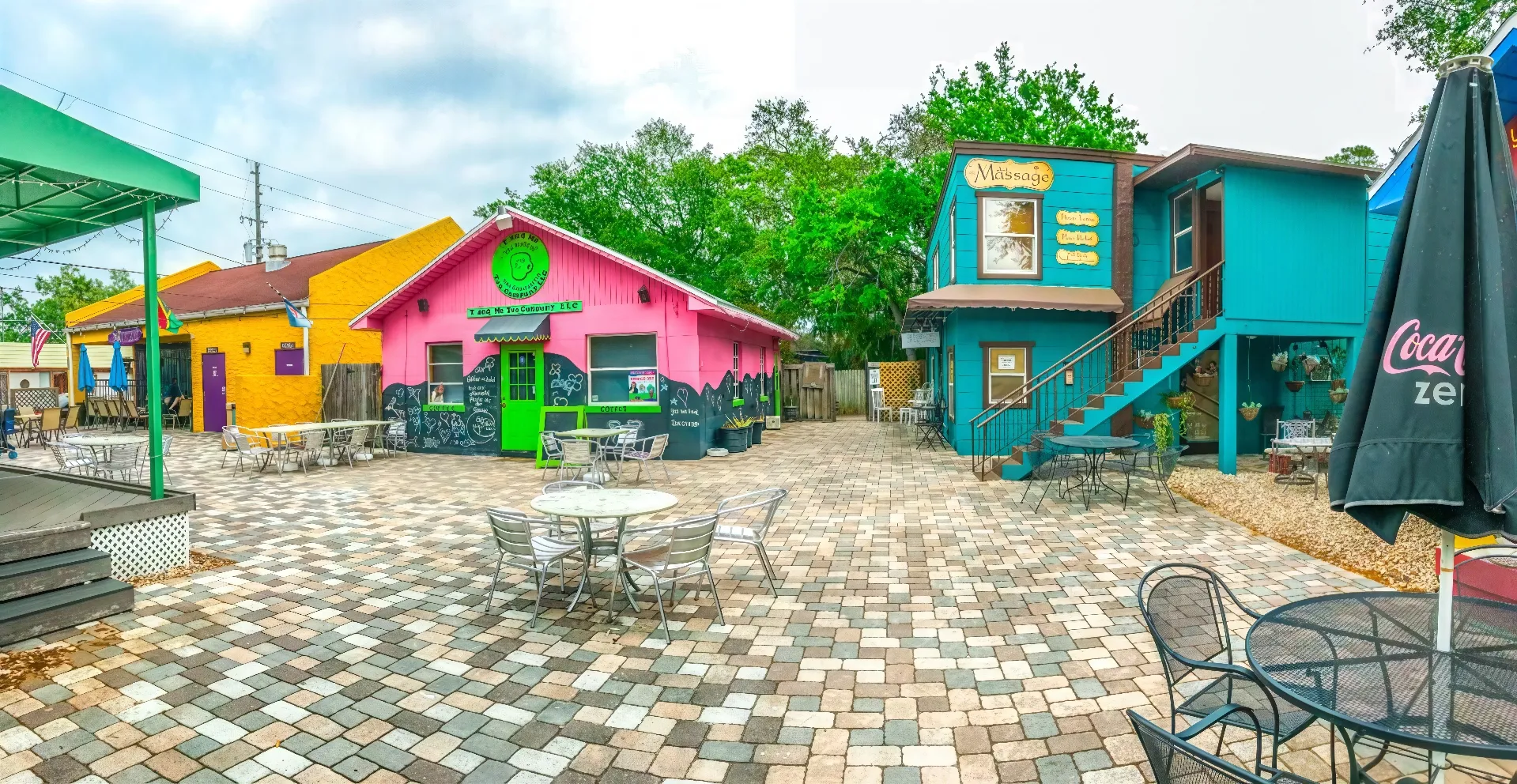 A colorful courtyard features a vibrant pink building with green accents, a teal building with stairs on the right, and outdoor seating on the cobblestone ground. Bright and inviting with lush greenery in the background