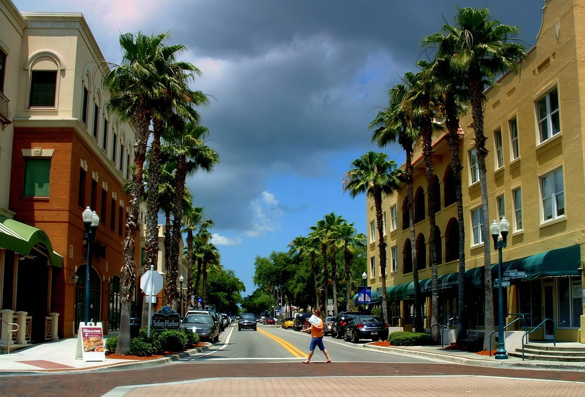 A person walks across a crosswalk on a palm tree-lined street in Safety Harbor, with tall buildings on either side. The sky is partly cloudy, with dark clouds contrasting the patches of blue.