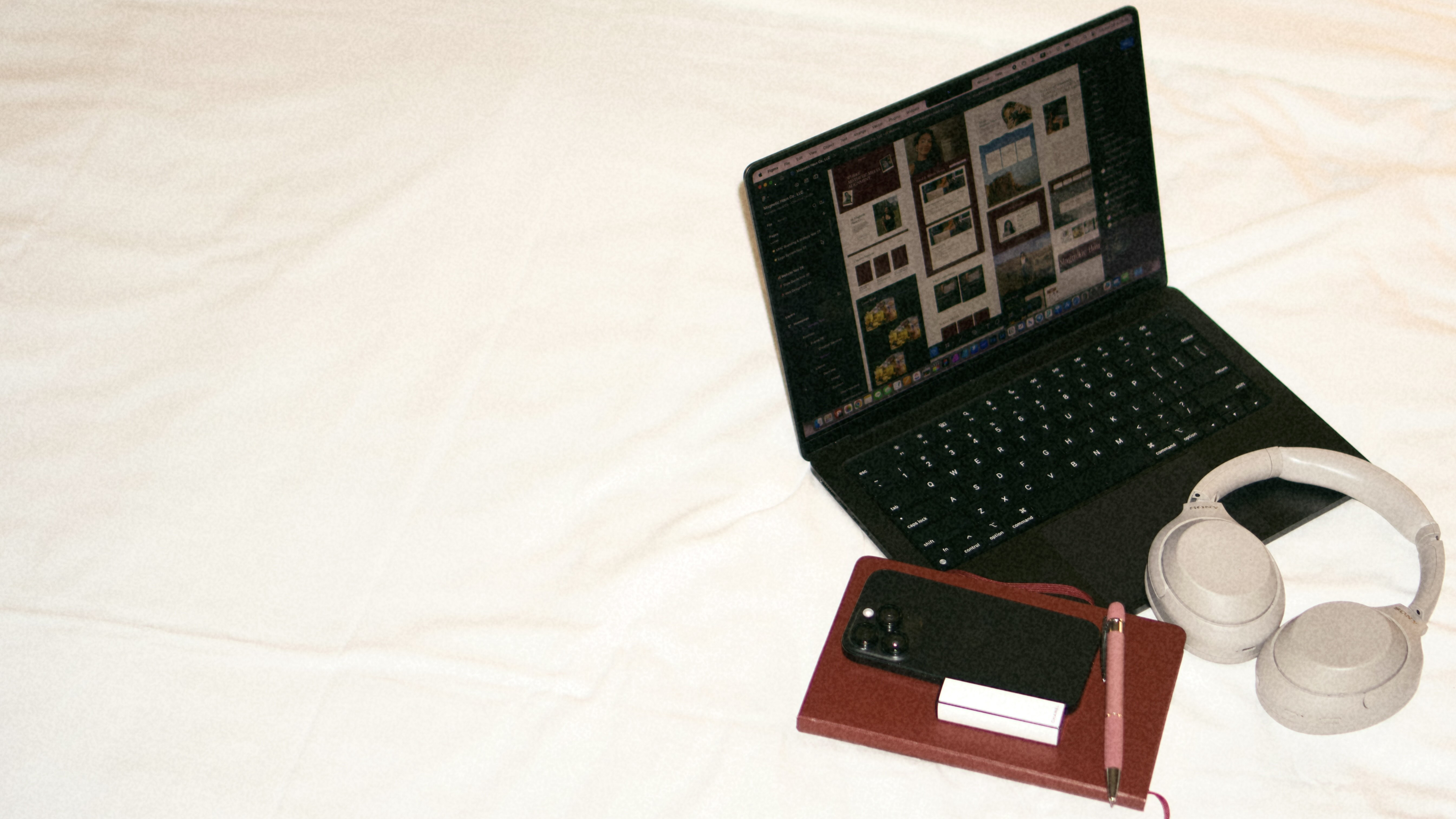 Open laptop displaying a design editing software next to white over-ear headphones, a red notebook, a black smartphone, a white charging case, and a pink pen on a white surface.