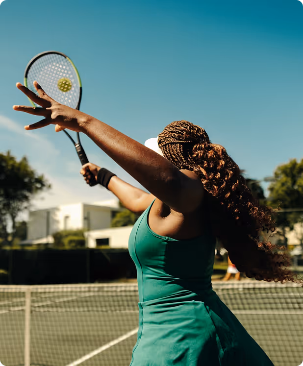 Woman in a green tennis dress serving on an outdoor court under a clear blue sky.