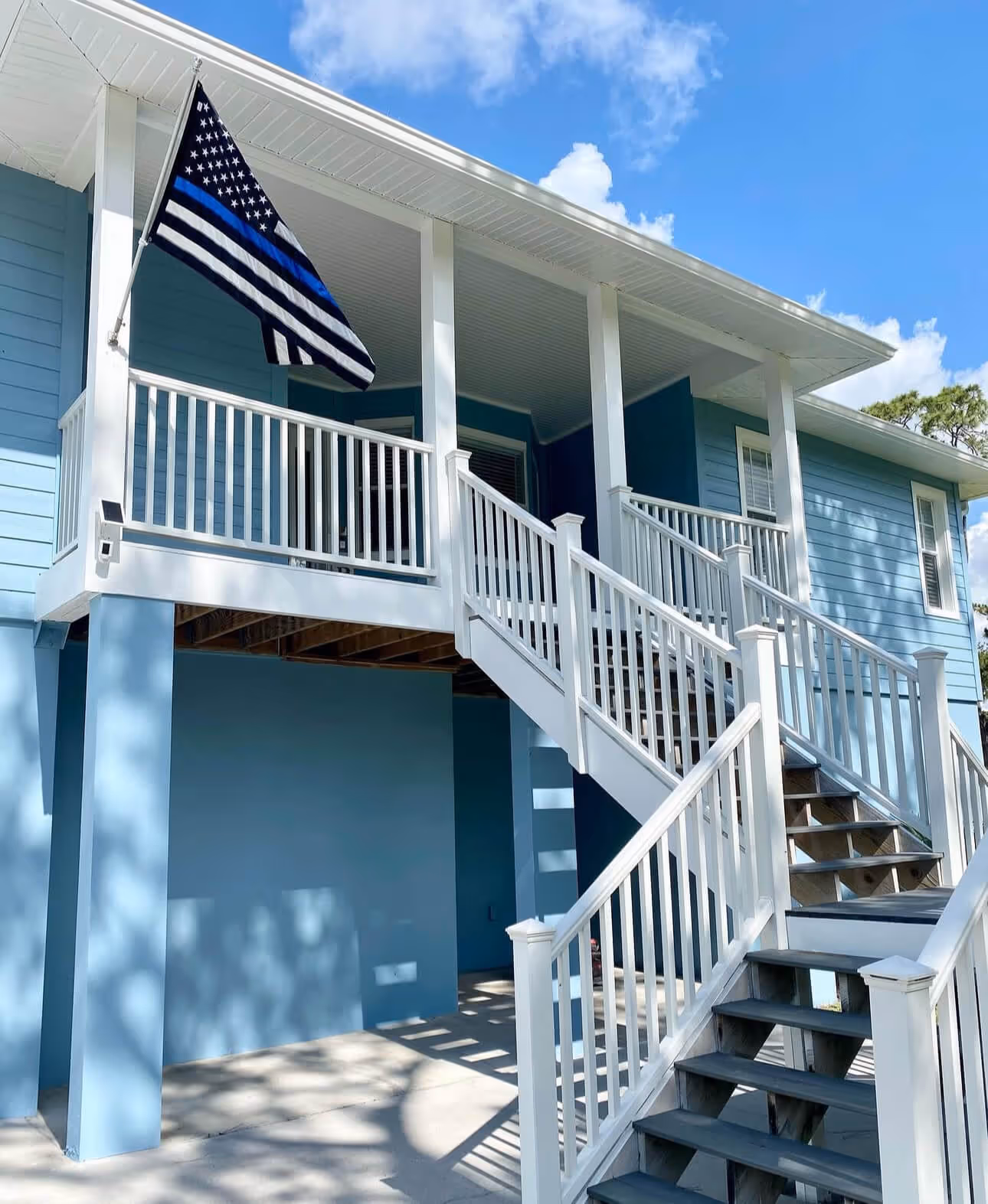 Light blue elevated house with white railings and stairs, flying a thin blue line American flag under blue sky.