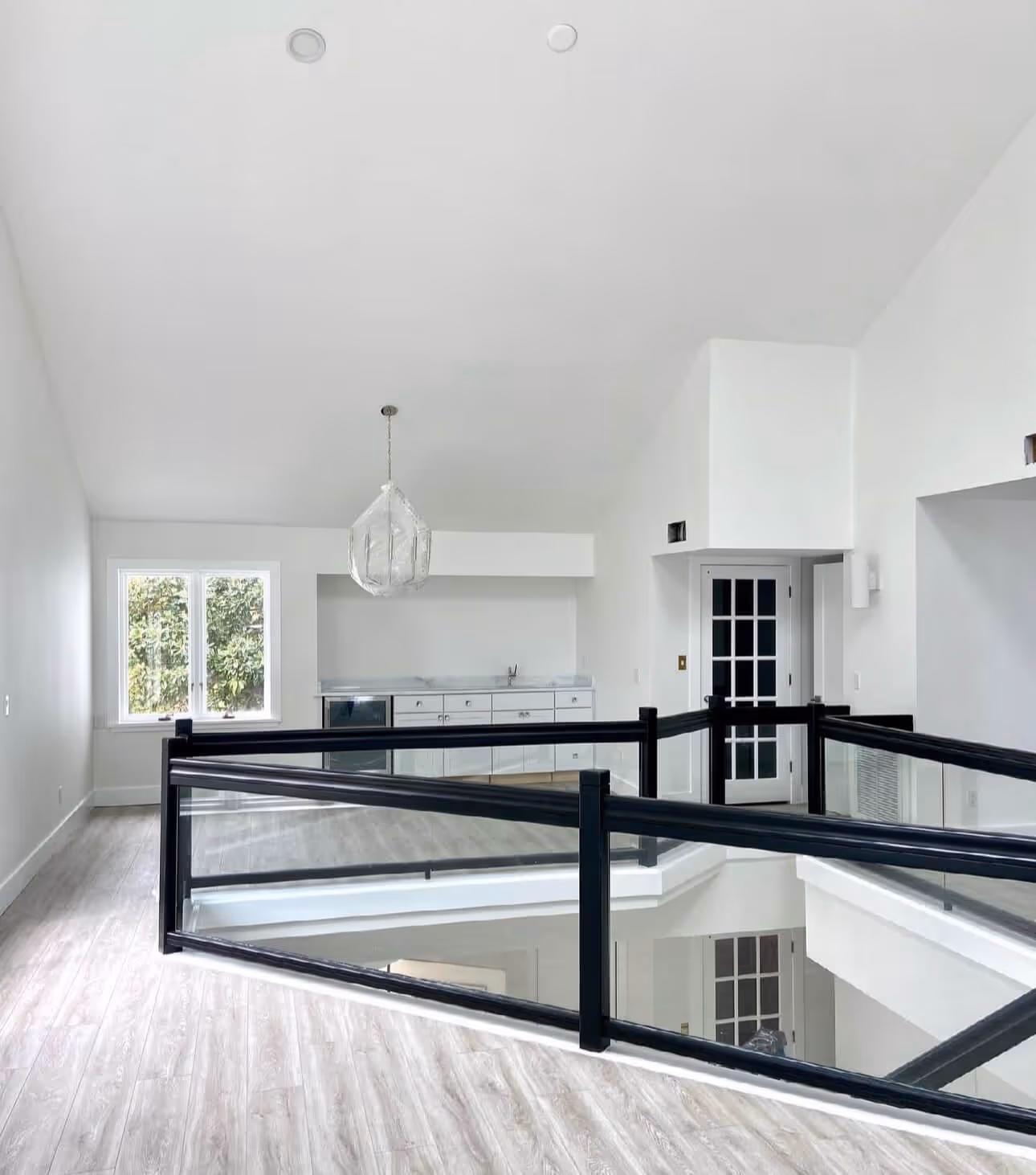 Bright modern upper-level room with light wood flooring, black metal and glass railing, white cabinetry, and a decorative hanging light fixture.