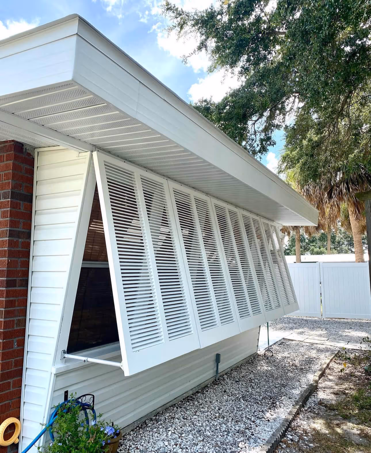 White exterior window with angled louvered shutters partially open on a white house with a brick corner, gravel path, and trees in the background.