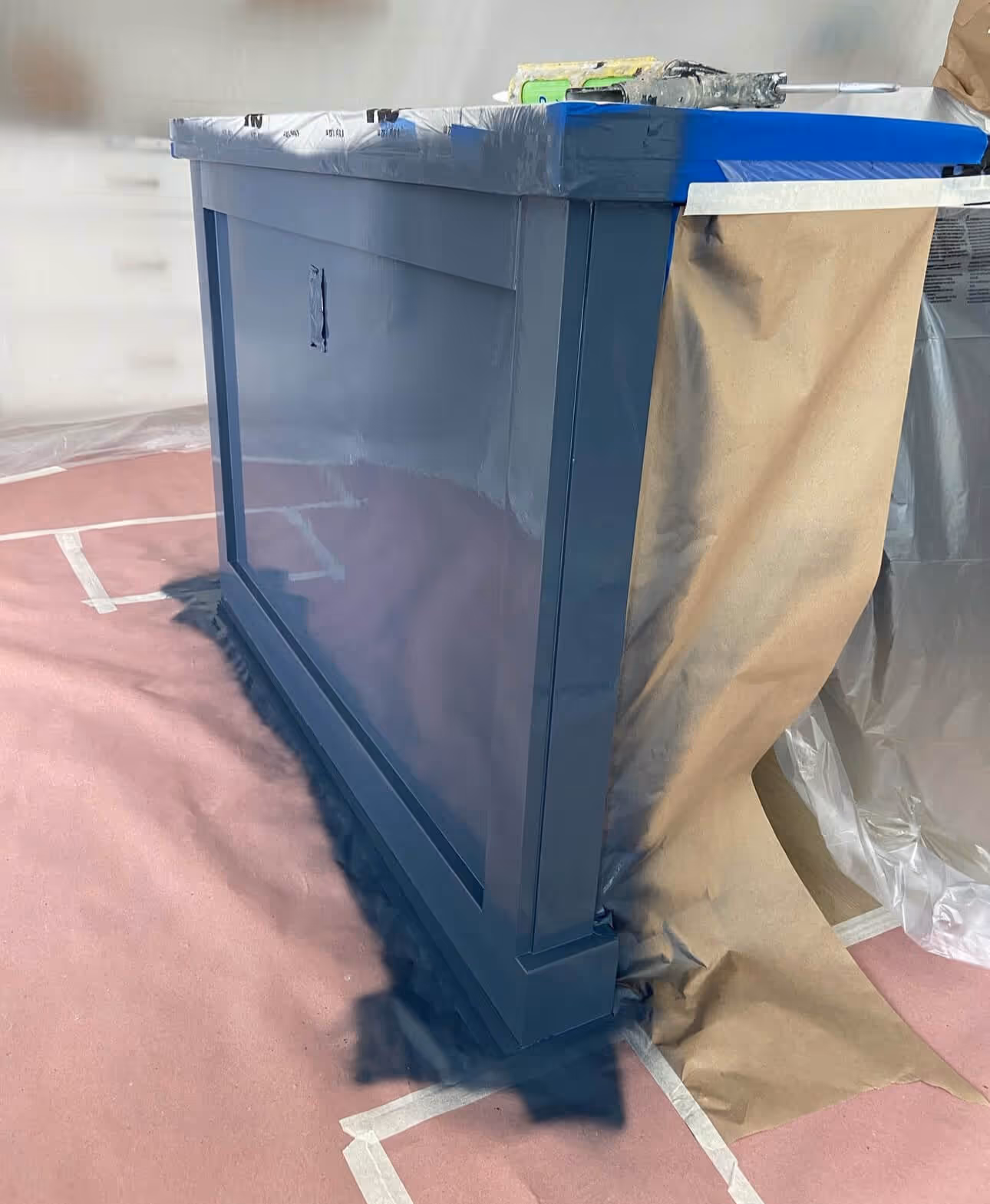 Blue painted cabinet with masking paper and tape for protection during painting on floor covered with protective paper.