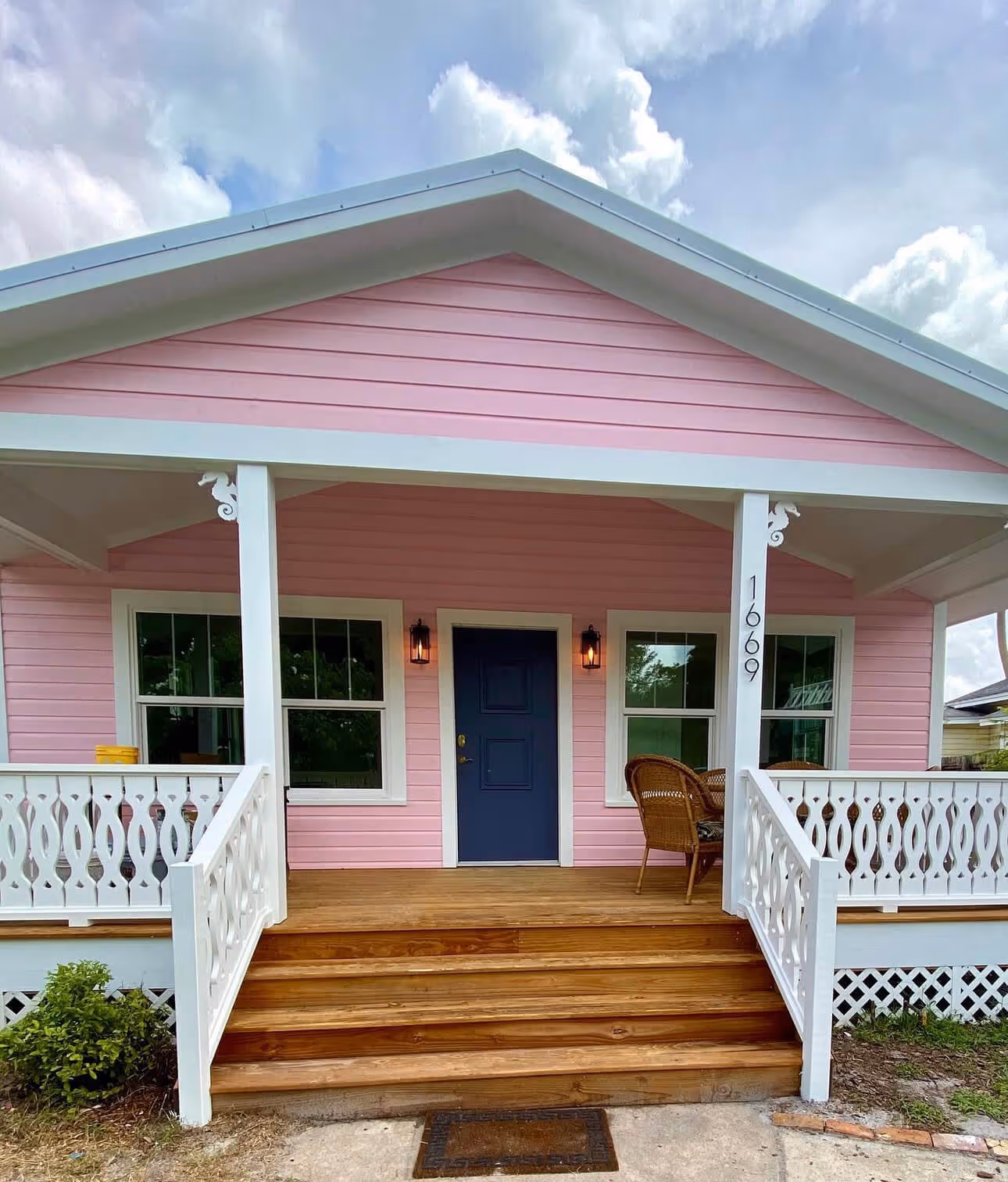 Front porch of a pink house with white railing, wooden steps, dark blue door, two windows, and wicker chairs on the right side.