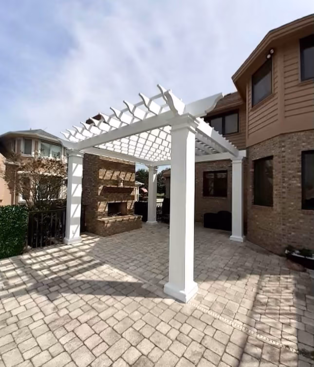 Outdoor patio area with a white pergola structure, stone fireplace, and paved stone flooring next to a brick house.