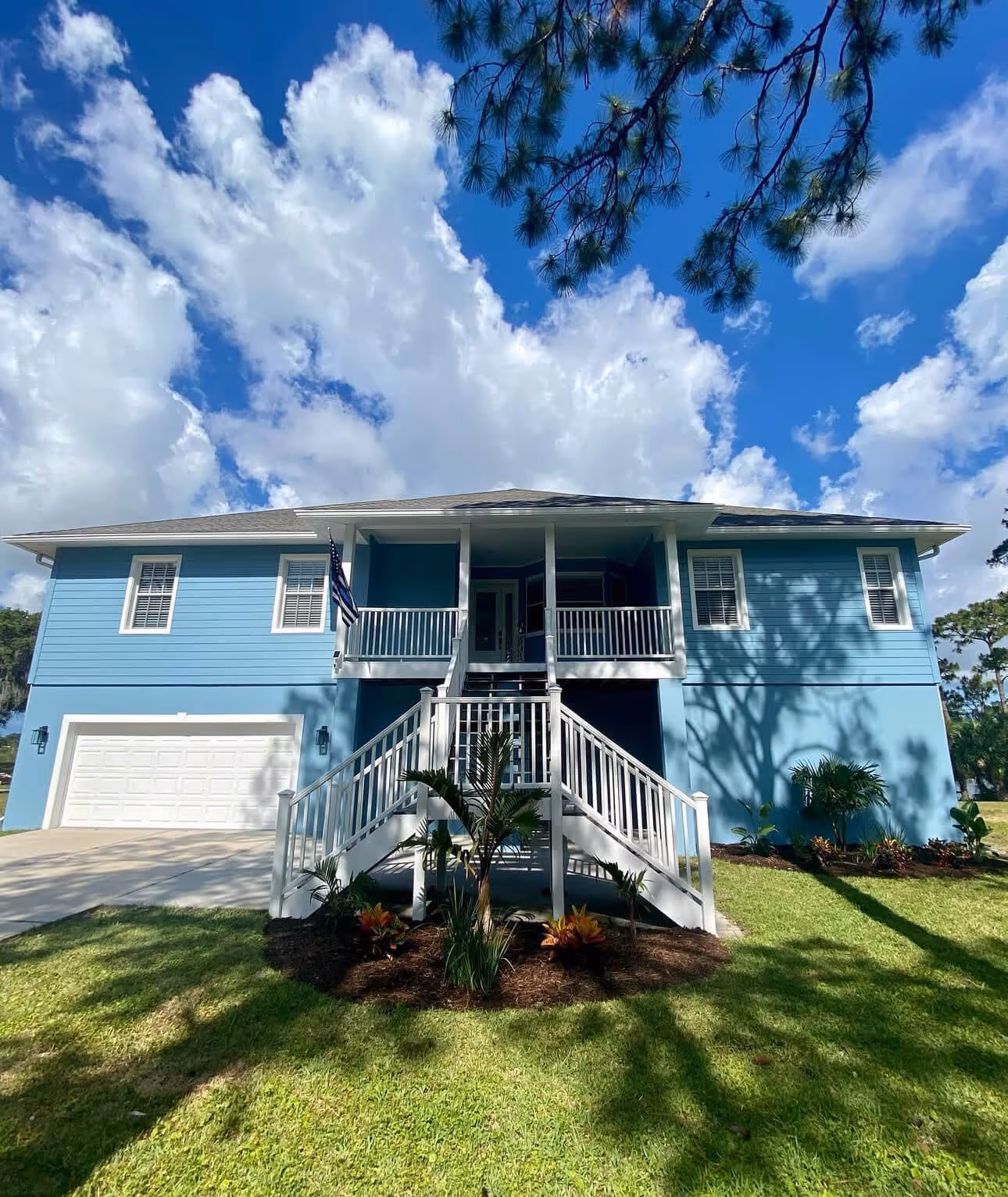 Two-story blue house with white railings, a front staircase, garage, and an American flag under a partly cloudy sky.