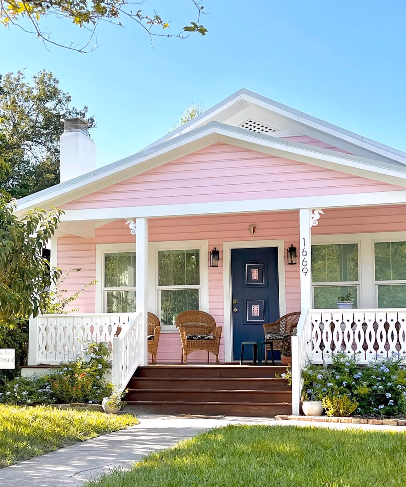 Front view of a small pink house with a dark blue door, white trim, a wooden porch with wicker chairs, and a green lawn.