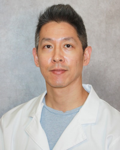 Man with short black hair wearing a white lab coat and light blue shirt against a gray background.