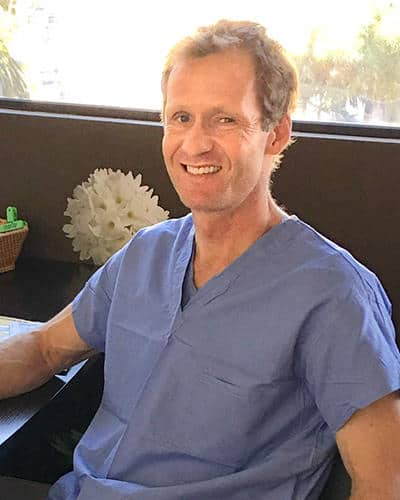 Smiling man wearing blue medical scrubs sitting at a desk with a decorative object in the background.