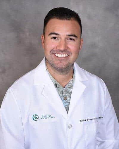 Smiling man with short dark hair wearing a white medical coat over a patterned shirt against a gray background.
