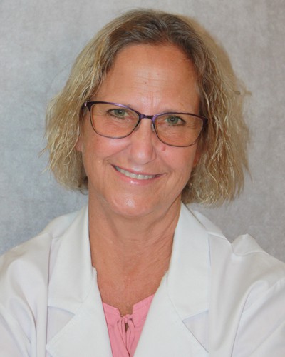 Smiling woman with blonde curly hair wearing glasses, a white lab coat, and a pink top in front of a gray background.