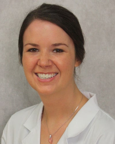 Smiling woman with dark hair wearing a white lab coat and a pink pendant necklace against a neutral background.