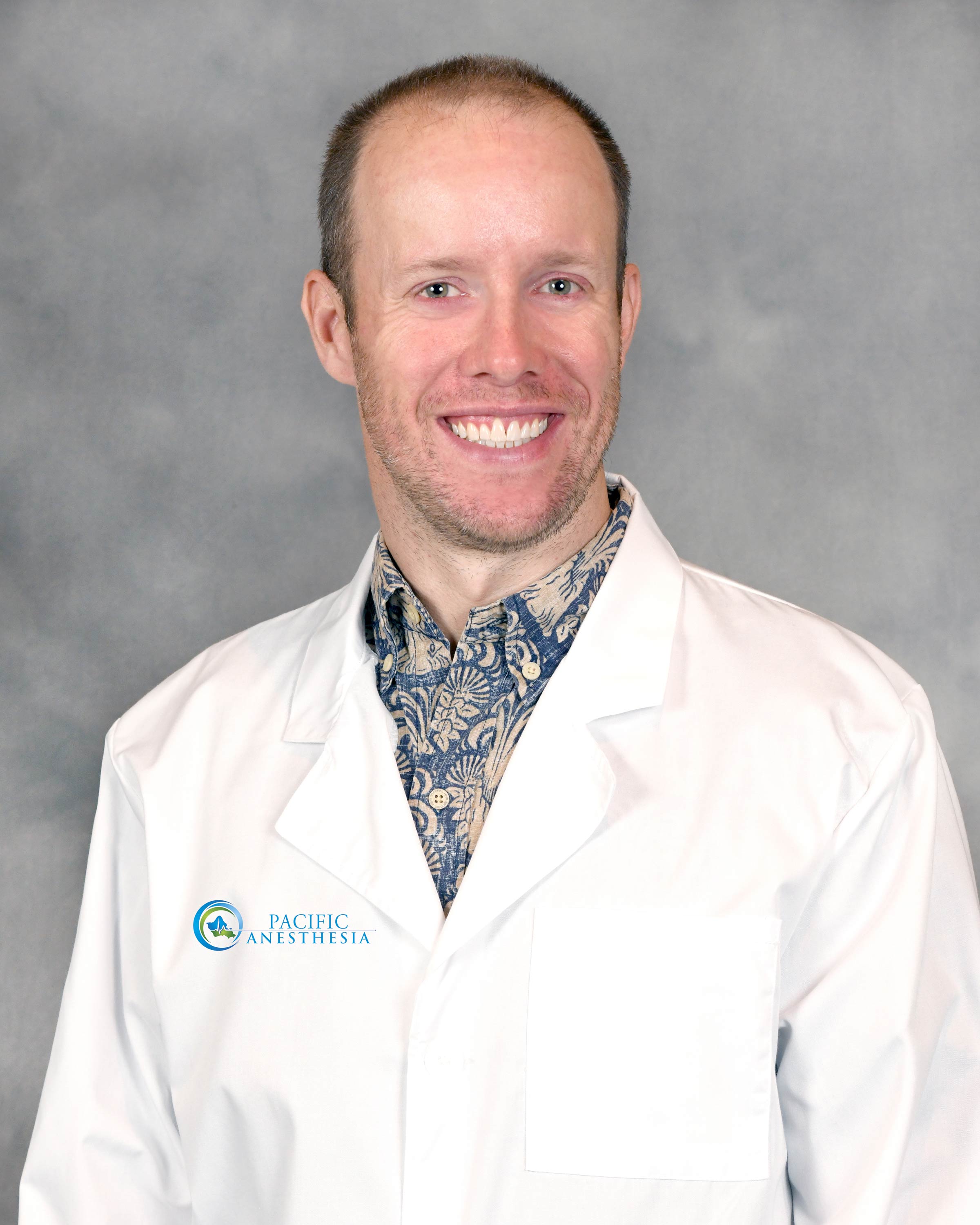 Smiling man wearing a patterned shirt under a white lab coat with Pacific Anesthesia logo on the chest.