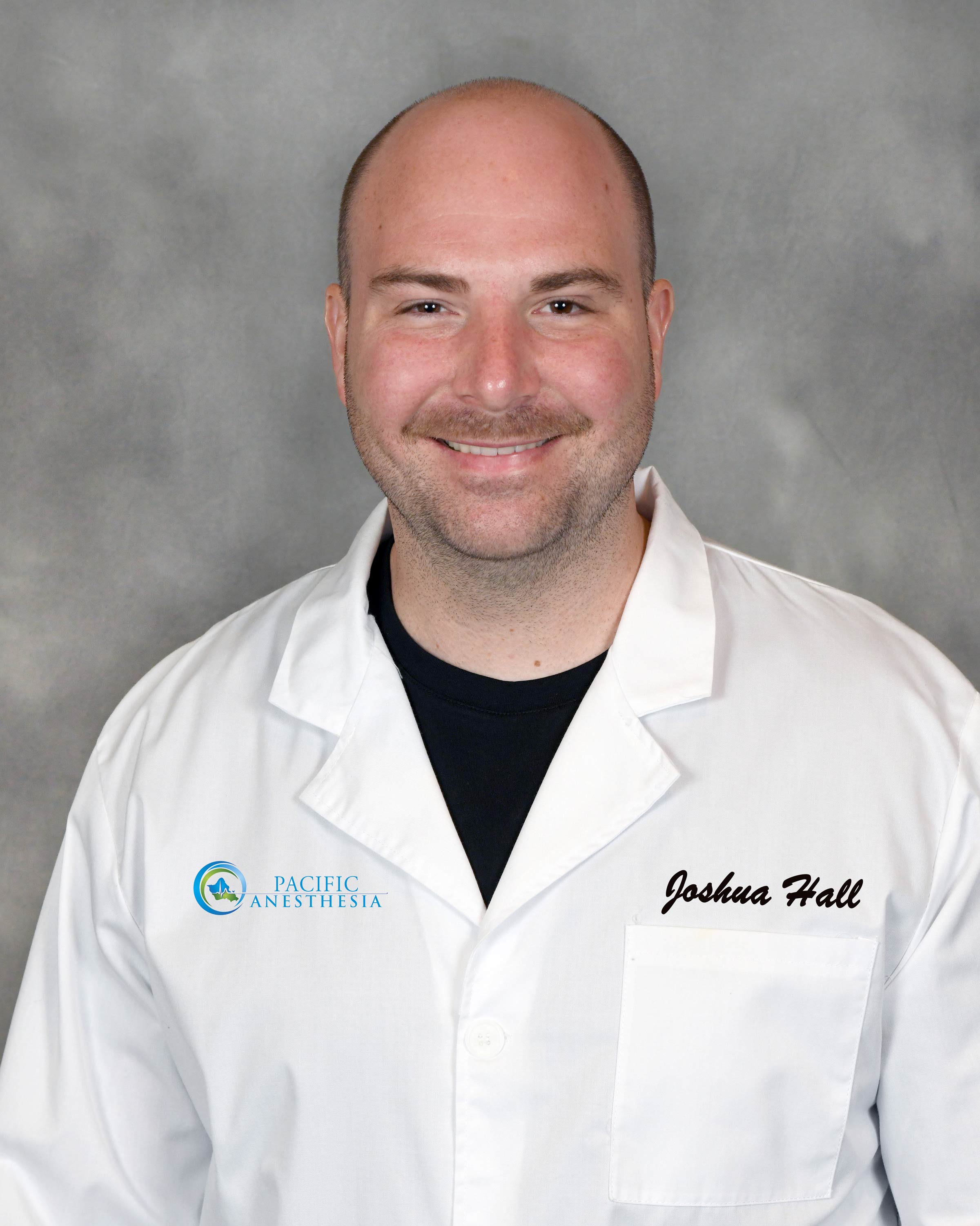 Smiling man wearing a white Pacific Anesthesia lab coat with the name Joshua Hall embroidered on it.