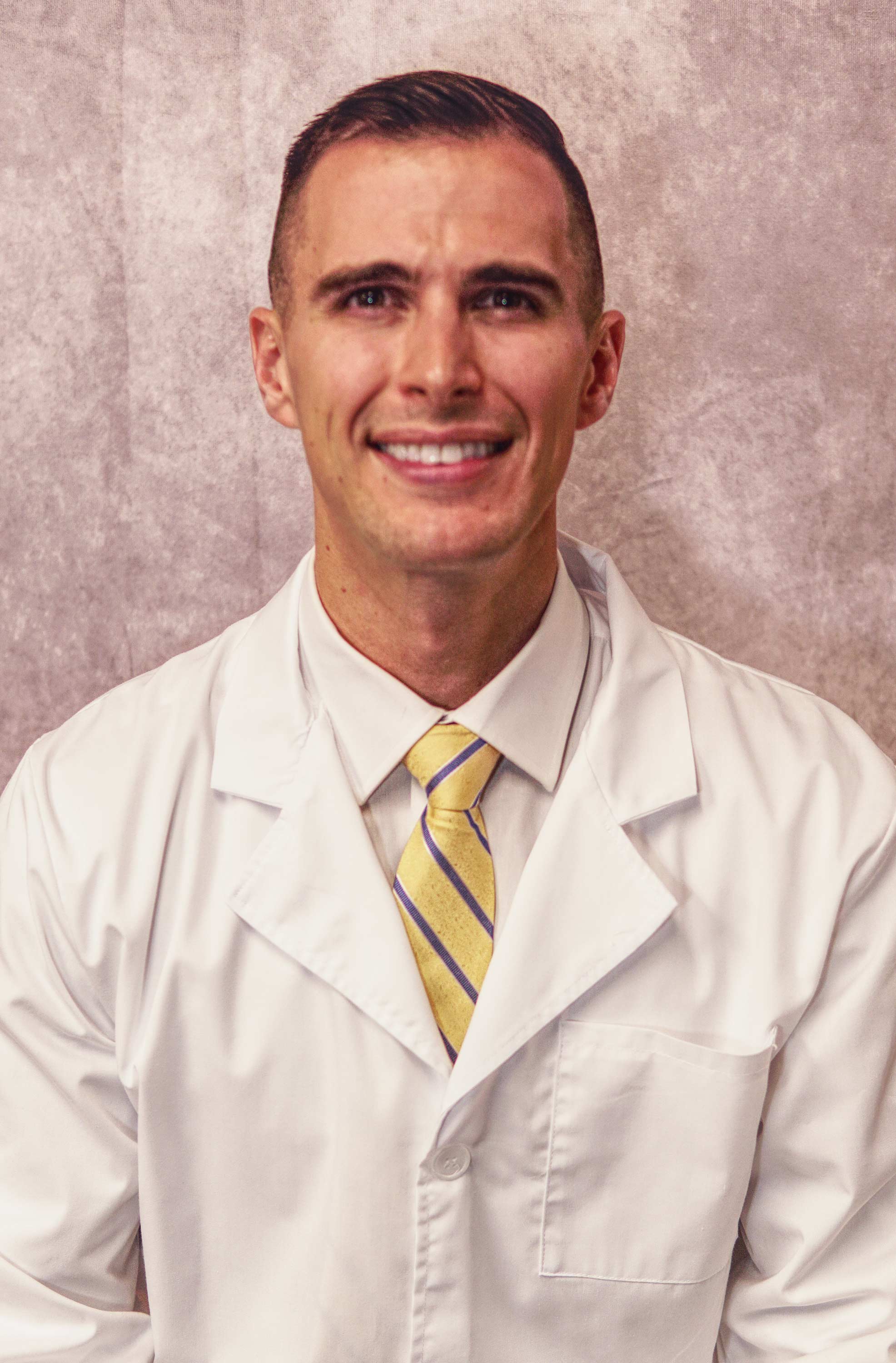 Smiling man wearing a white lab coat, yellow striped tie, and white shirt against a neutral background.