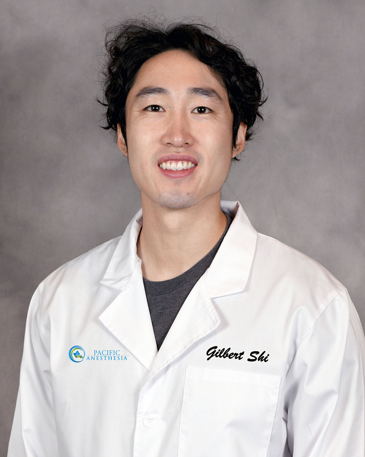 Headshot of a smiling man with dark curly hair wearing a white lab coat with 'Gilbert Shi' embroidered and a Pacific Anesthesia logo.