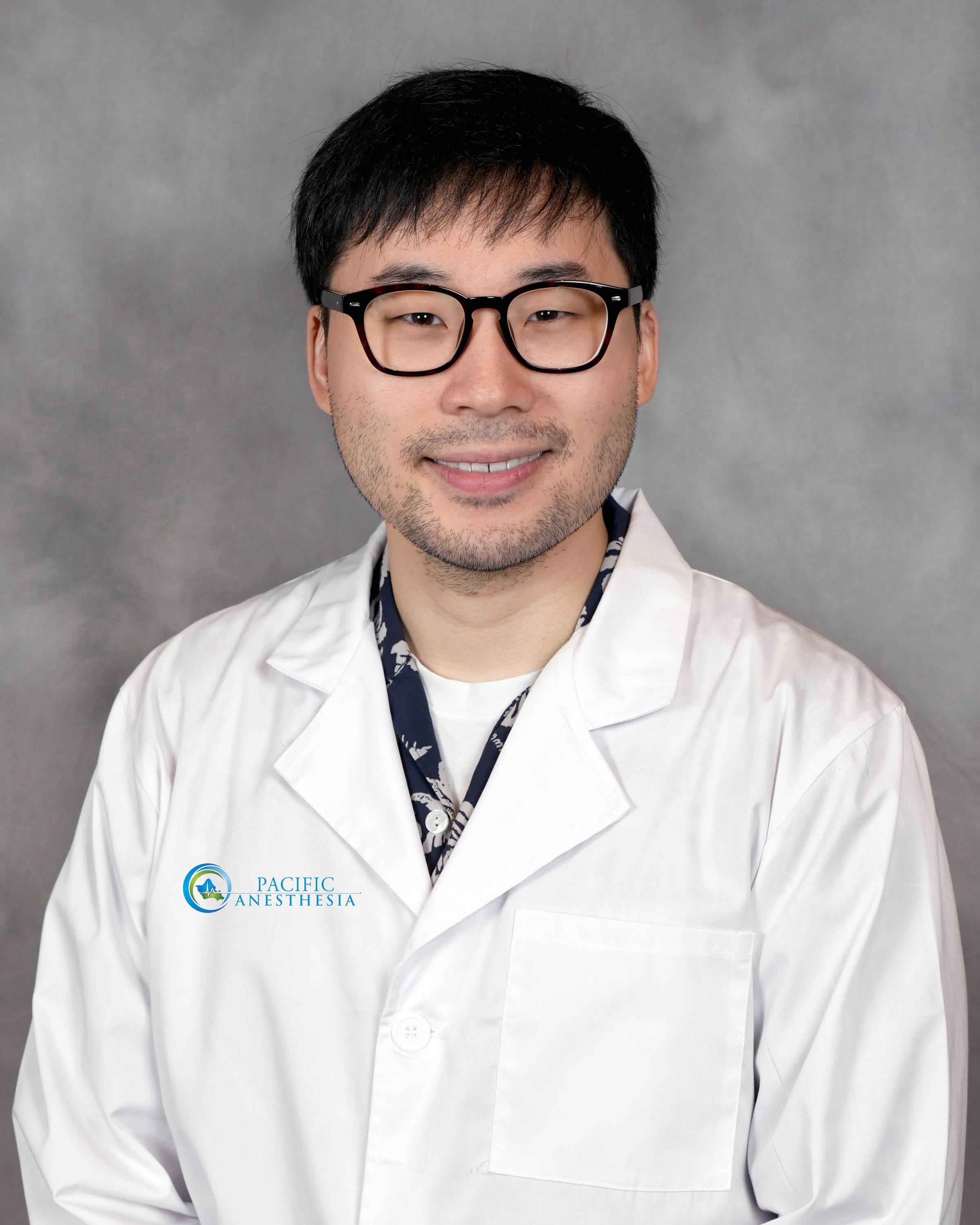 Smiling man wearing glasses and a white lab coat with Pacific Anesthesia logo on a gray background.