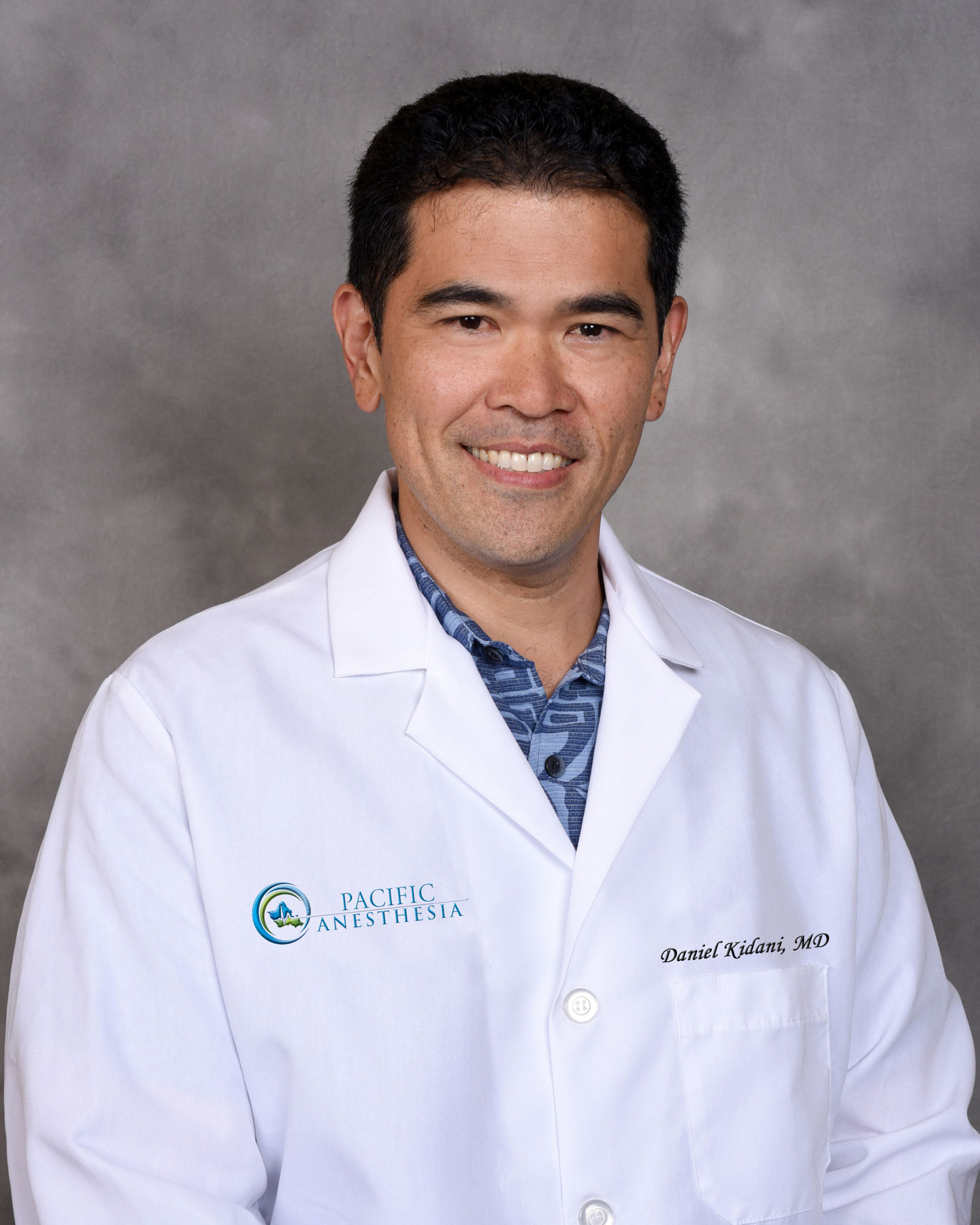 Man smiling wearing a white lab coat with Pacific Anesthesia logo and name Daniel Kidani, MD.