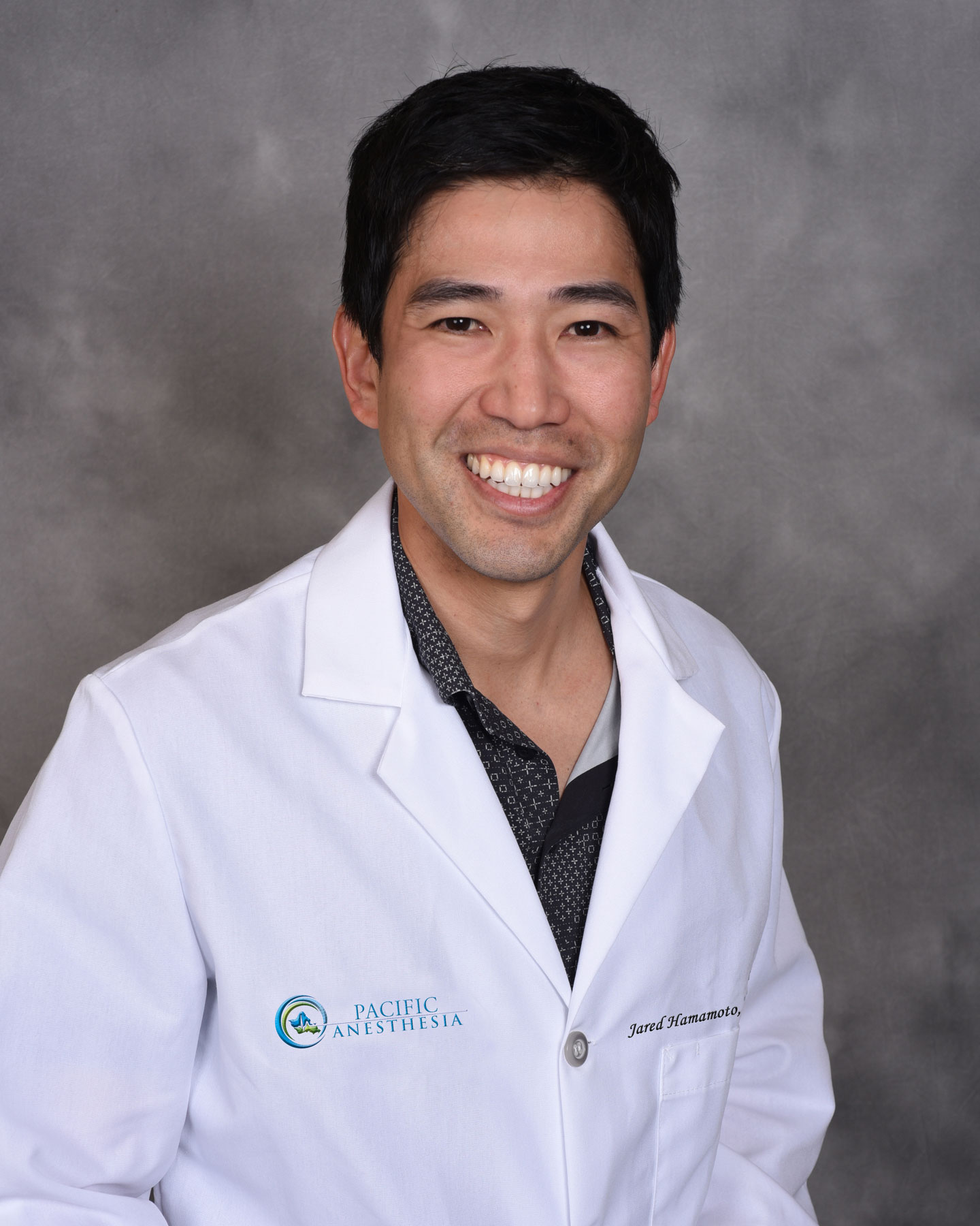 Smiling man in a white medical coat with Pacific Anesthesia logo and name embroidered, against a gray backdrop.