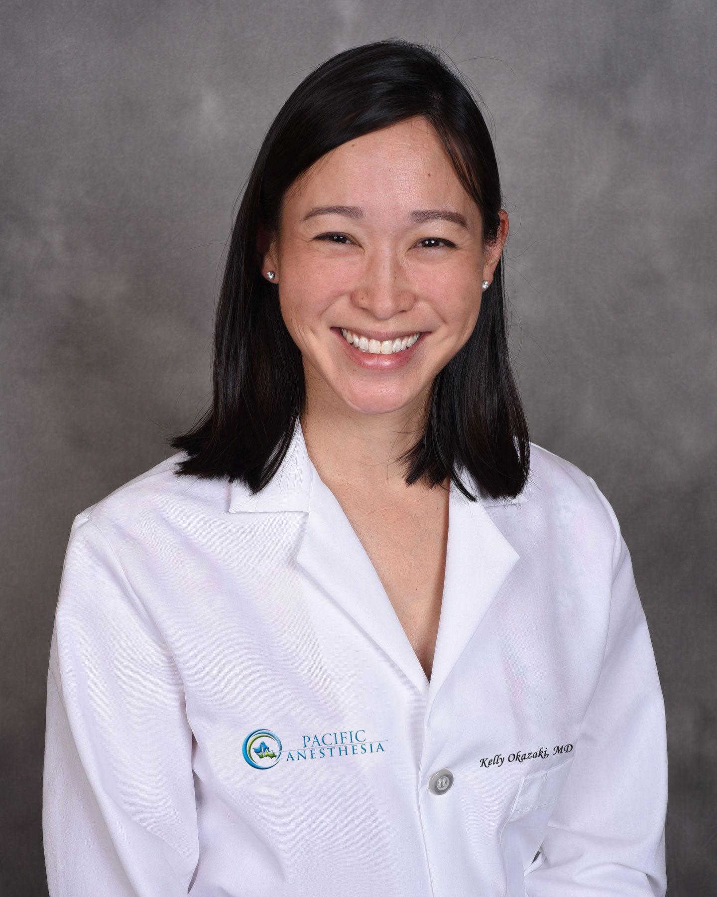 Smiling woman with shoulder-length black hair wearing a white lab coat with 'Pacific Anesthesia' and 'Kelly Okazaki, MD' embroidered on it.