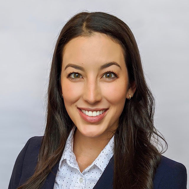 Smiling woman with long dark hair wearing a navy blazer and white polka dot blouse against a light gray background.