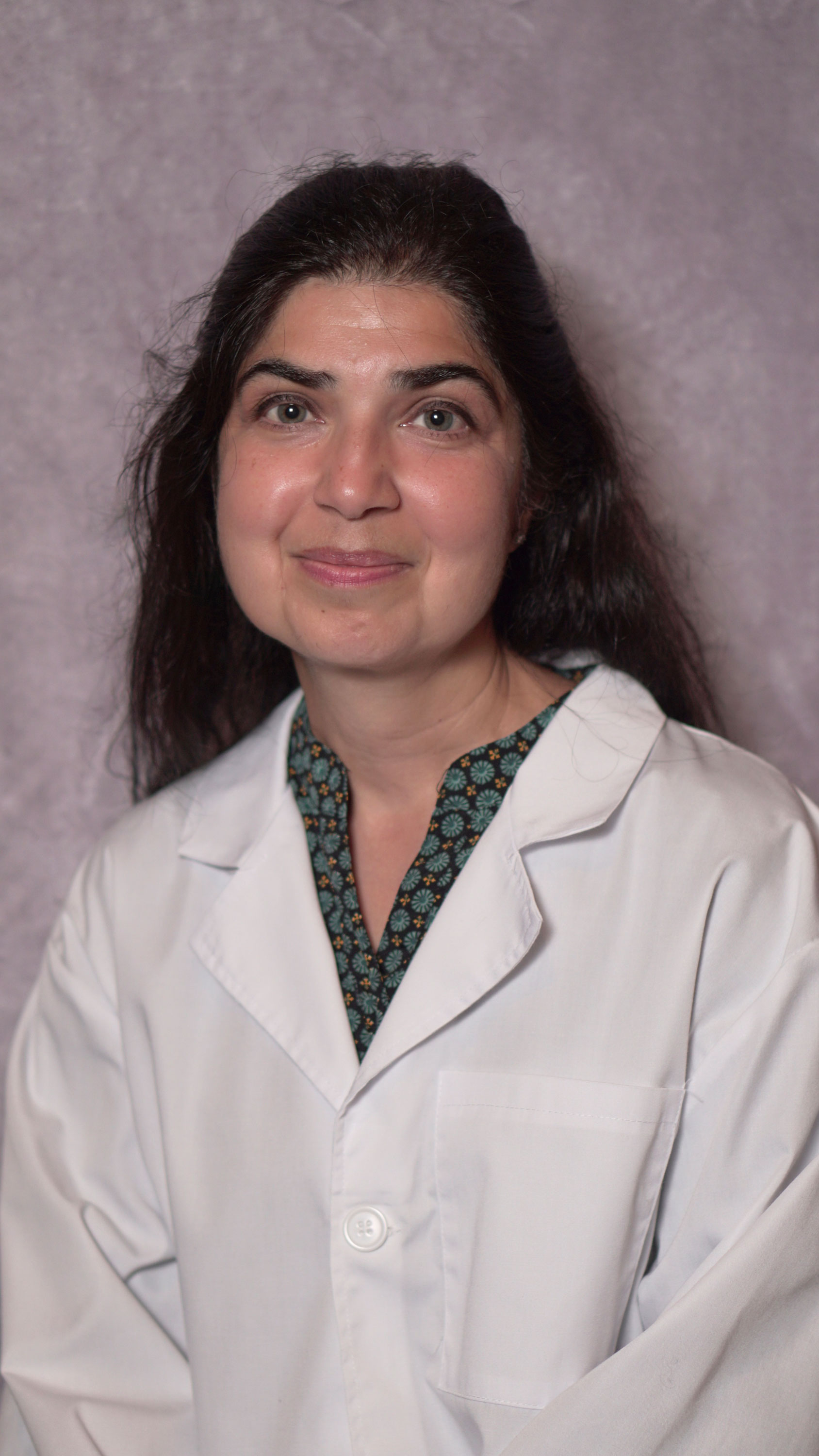Smiling woman with long dark hair wearing a white lab coat over a patterned green blouse.