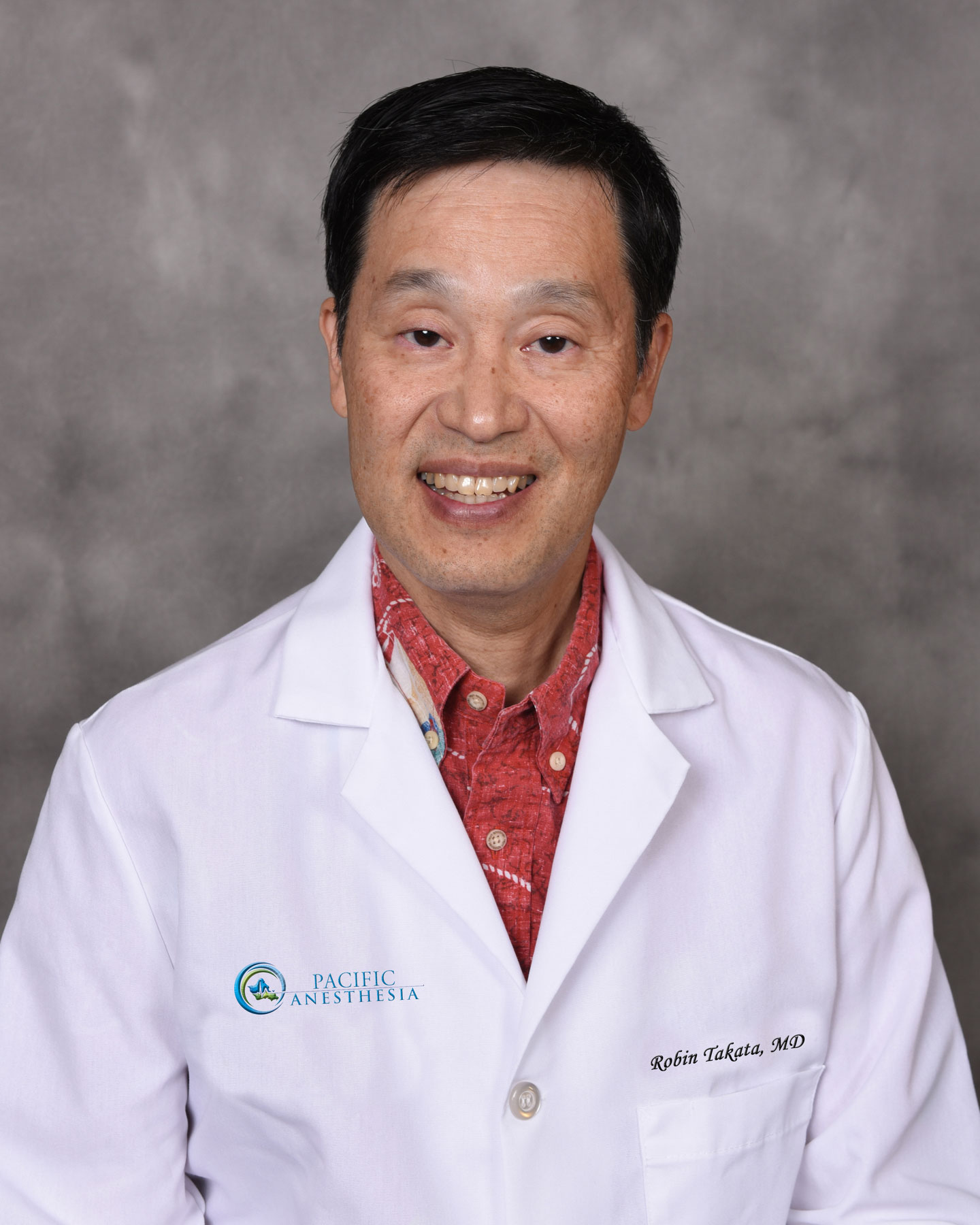 Smiling man wearing a white medical coat with Pacific Anesthesia logo and a red shirt underneath.