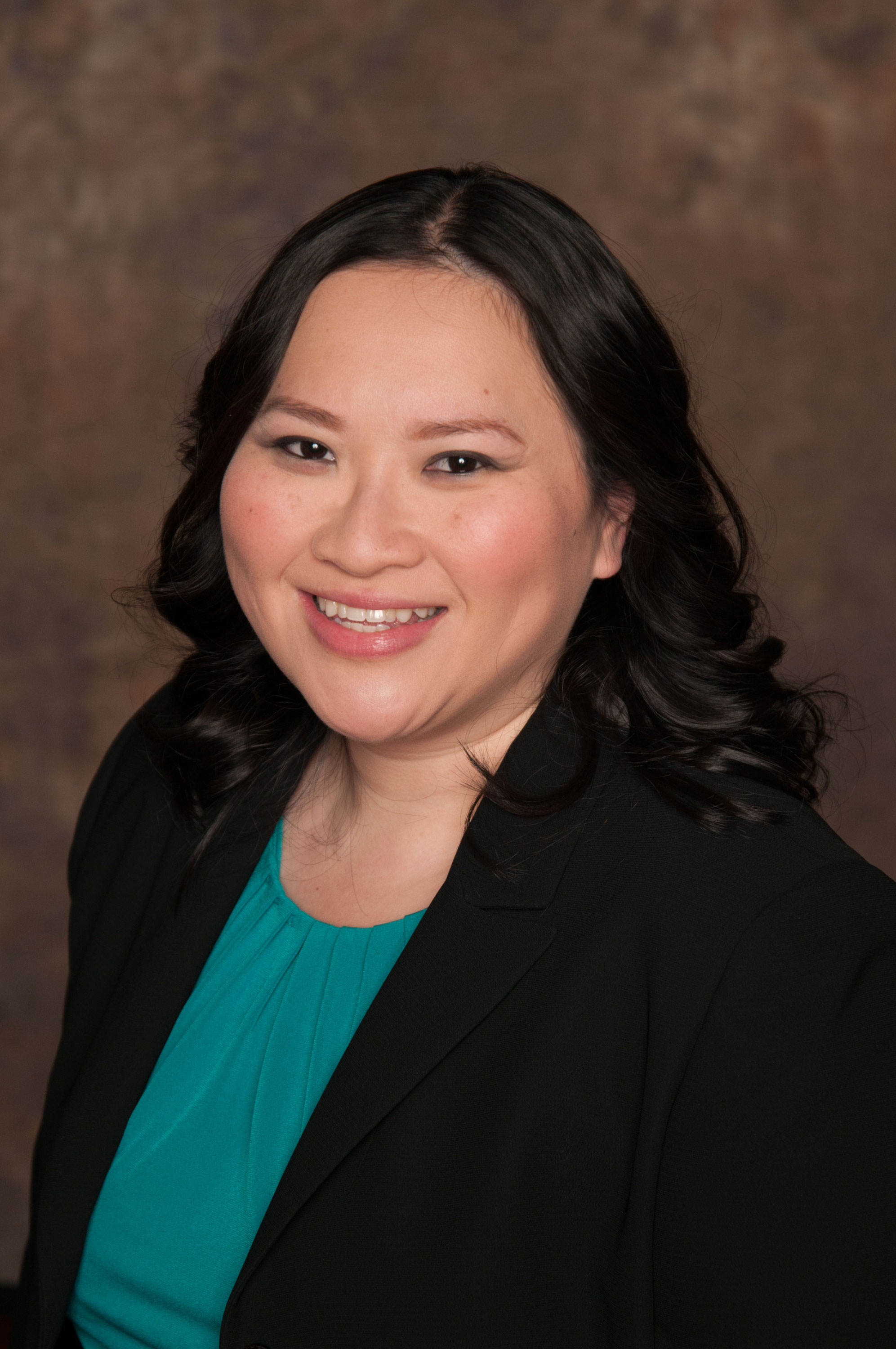 Smiling woman with wavy black hair wearing a teal blouse and black blazer against a brown background.