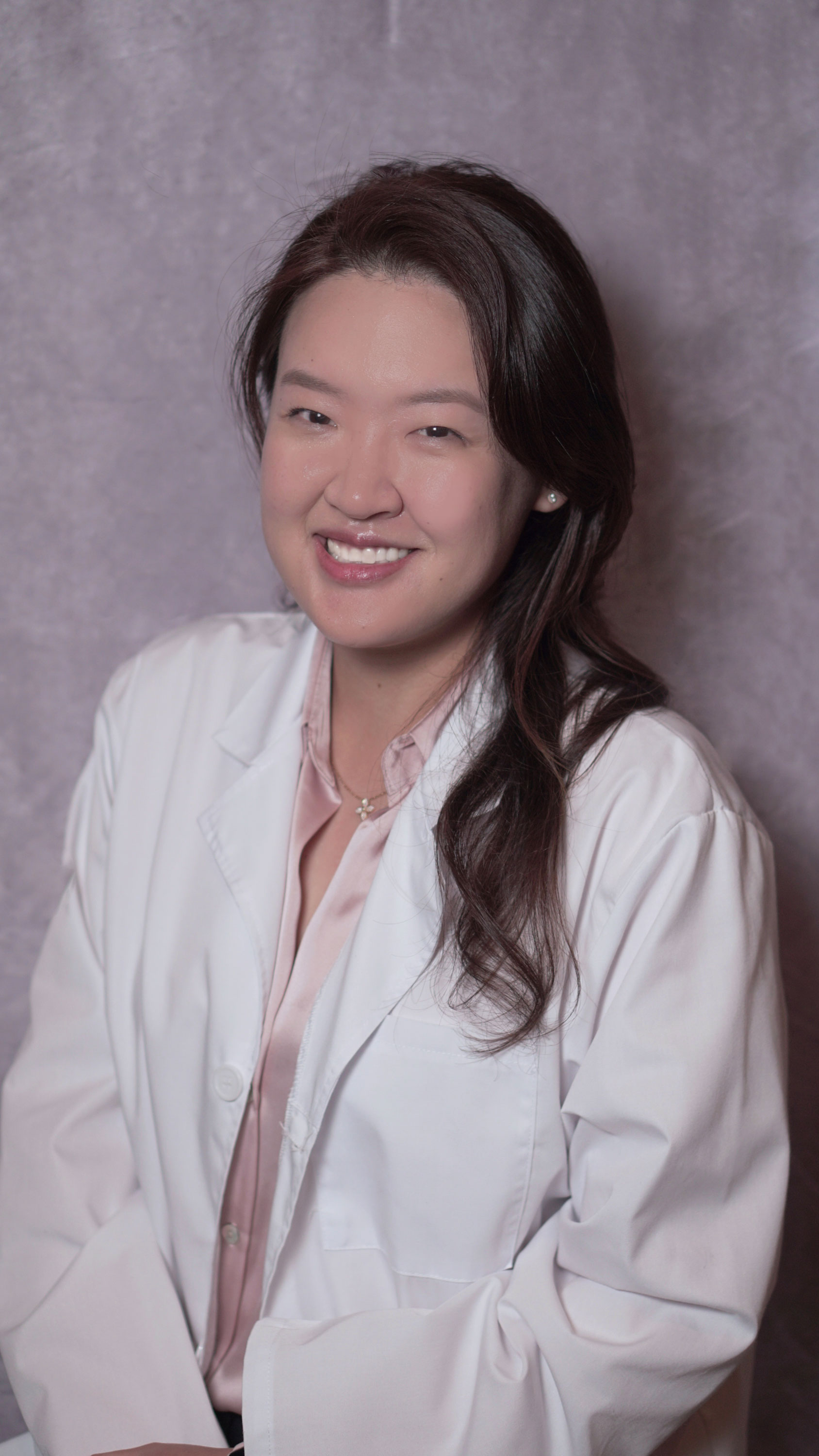 Smiling woman with long dark hair wearing a white lab coat and pink blouse against a gray backdrop.