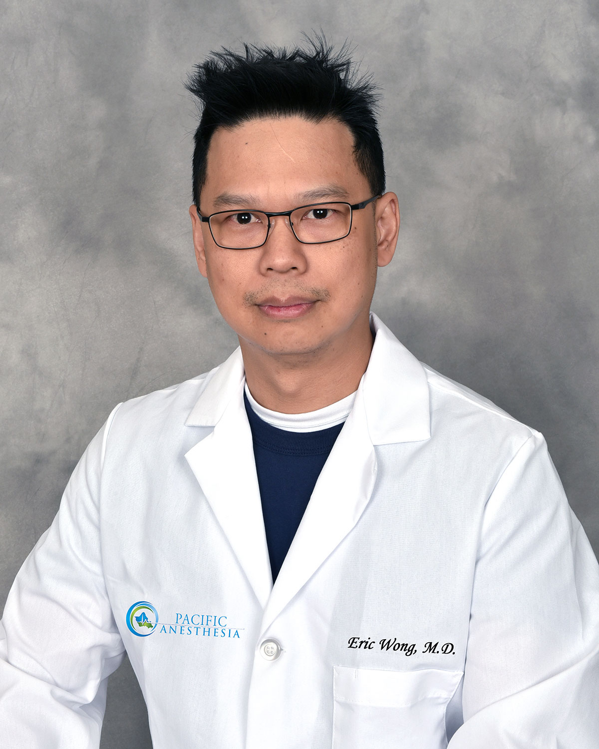 Male doctor wearing glasses and a white lab coat with 'Pacific Anesthesia' logo and 'Eric Wong, M.D.' embroidered on it.