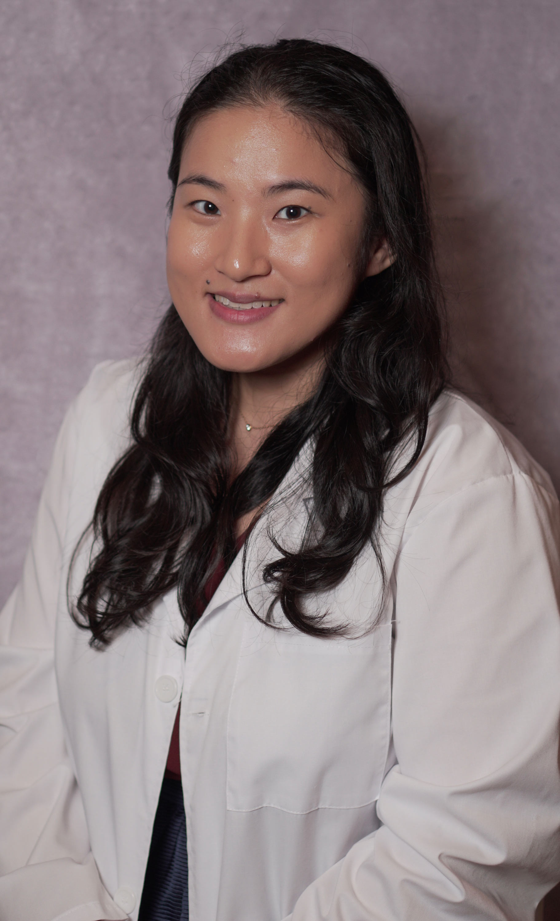 Smiling woman with long dark hair wearing a white lab coat against a soft purple background.