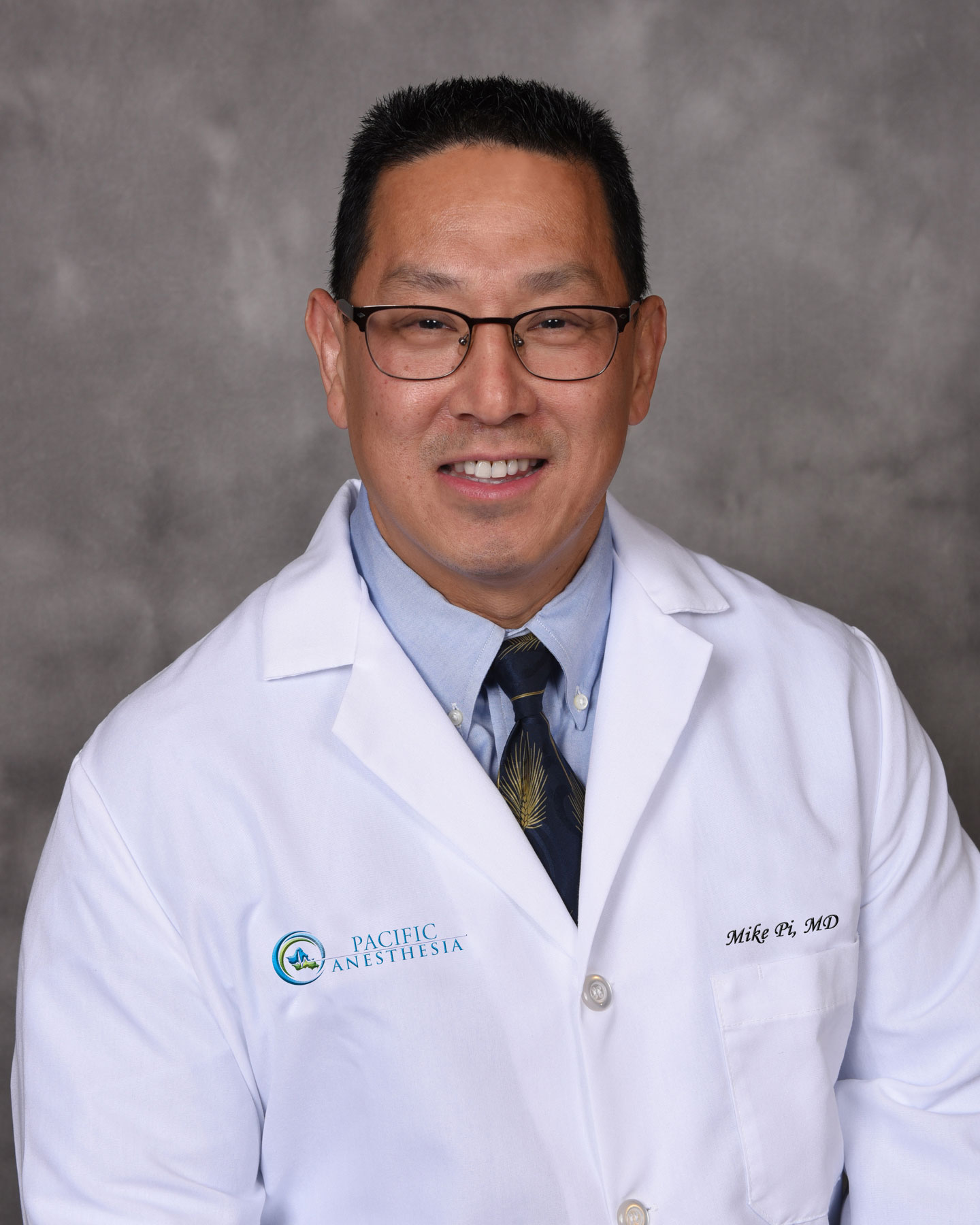 Smiling male doctor with glasses wearing a white coat, blue shirt, and patterned tie against a gray background.